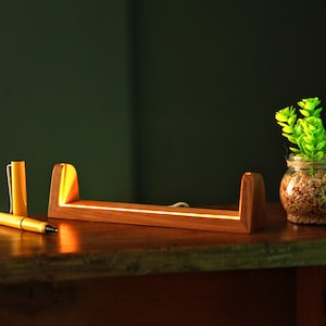 Puede incluir: Un organizador de escritorio de madera con una luz LED integrada, un bol&iacute;grafo amarillo y un peque&ntilde;o tarro de cristal con una planta verde. El organizador de escritorio es de madera y tiene un brillo c&aacute;lido.