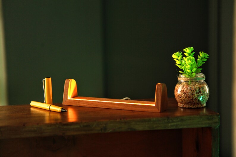 Puede incluir: Un organizador de escritorio de madera con una luz LED integrada, un bol&iacute;grafo amarillo y un peque&ntilde;o tarro de cristal con una planta verde. El organizador de escritorio es de madera y tiene un brillo c&aacute;lido.