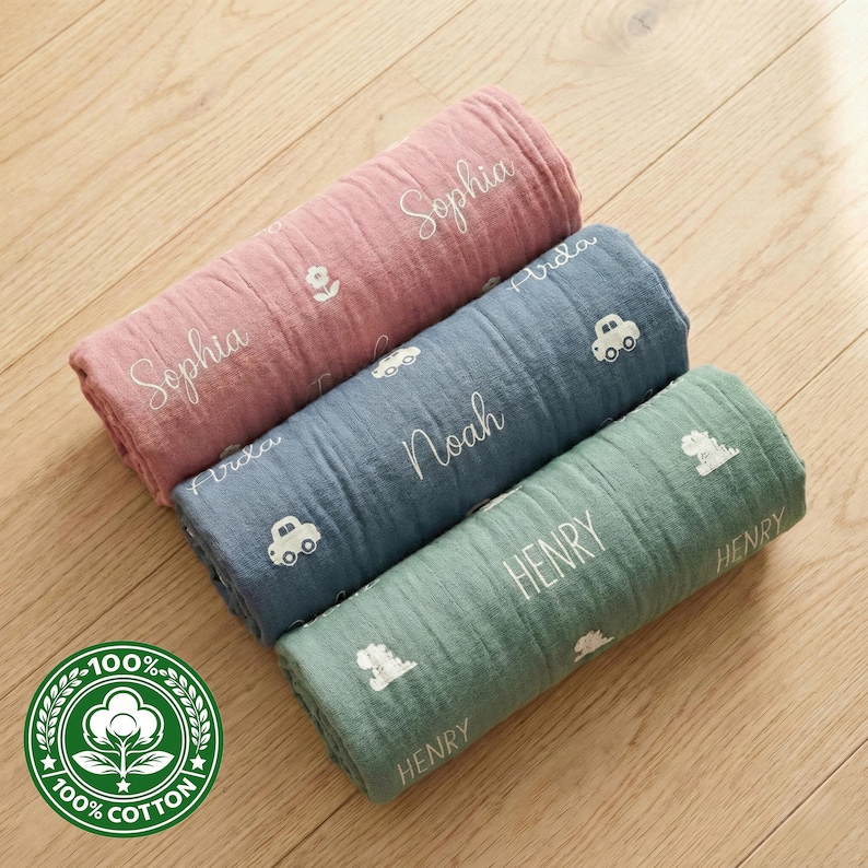 May include: Three rolled cotton baby blankets in pink, blue, and green. Each blanket is embroidered with a name and a small graphic. The pink blanket has the name "Sophia" and a flower. The blue blanket has the name "Noah" and car graphics. The green blanket has the name "Henry" and a dinosaur graphic.