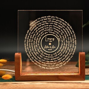 May include: A wooden base with a clear acrylic panel featuring a circular design with a gold inscription. The inscription reads "Whenever I'm alone with you You make me feel like I am young again Whenever I'm alone with you You make me feel like I am whole again Whenever I'm alone with you I will always love you However long I stay I will always love you However far away I will always love you Whatever words I say I will always love you 'Cause I love you". The centre of the circle features a smaller circle with the text "Lovesong" and "A&Am".