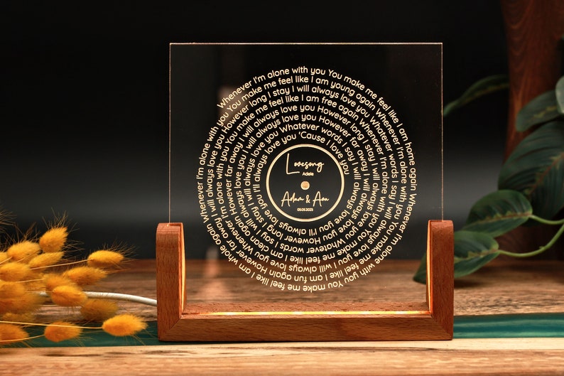 May include: A wooden base with a clear acrylic panel featuring a circular design with a gold inscription. The inscription reads "Whenever I'm alone with you You make me feel like I am young again Whenever I'm alone with you You make me feel like I am whole again Whenever I'm alone with you I will always love you However long I stay I will always love you However far away I will always love you Whatever words I say I will always love you 'Cause I love you". The centre of the circle features a smaller circle with the text "Lovesong" and "A&Am".