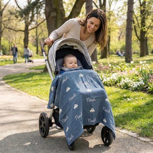 May include: A baby in a stroller is covered with a blue blanket. The blanket has white embroidered cars and the name "Noah". A woman is pushing the stroller on a paved path in a park.