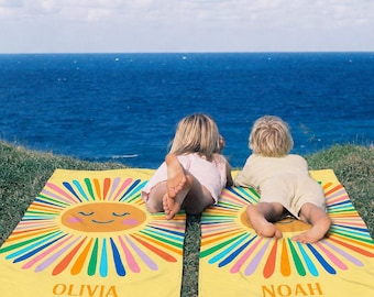 May include: Two rectangular yellow beach towels featuring a large, smiling sun graphic with multicolored rays in shades of pink, orange, blue, and green. The left towel is personalized with the name OLIVIA and the right with the name NOAH.
