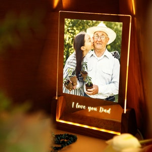 Puede incluir: Un marco de fotos de madera iluminado con una foto de una mujer besando a un hombre mayor. El marco tiene las palabras "I love you Dad" iluminadas en la base. La foto muestra al hombre con un sombrero y sosteniendo plantas.