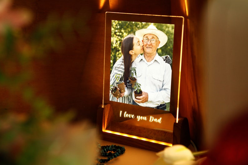 Puede incluir: Un marco de fotos de madera iluminado con una foto de una mujer besando a un hombre mayor. El marco tiene las palabras "I love you Dad" iluminadas en la base. La foto muestra al hombre con un sombrero y sosteniendo plantas.
