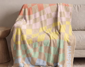 May include: A soft throw blanket featuring a wavy, distorted checkerboard pattern in pastel shades of peach, pink, yellow, mint green, and light blue against a tan background. The name HARPER is printed vertically in light blue capital letters.