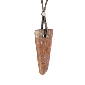 Puede incluir: Collar con colgante de piedra natural. El colgante es de color marrón apagado con motas rojas y grises, y un agujero para el cordón. El cordón es de color marrón oscuro y se sujeta con un anillo metálico plateado.