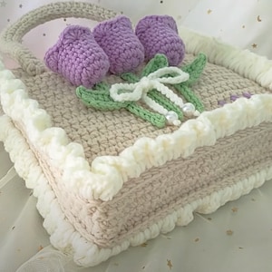 May include: A crocheted beige and white tissue box cover with a flower design. The flowers are purple and green and have a white bow with pearl accents.