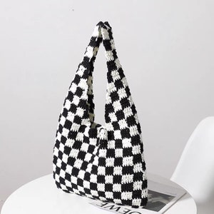 May include: A black and white chequered crochet tote bag. The bag has a wide, open top and two long handles. The bag is made of a soft, textured material and is sitting on a white surface.