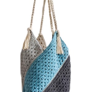 May include: A crocheted tote bag with a blue, grey, and beige colour scheme. The bag has a long, braided strap and a tassel on each side.