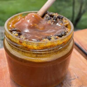 Op de afbeelding: Een heldere glazen pot gevuld met een donker amberkleurige vloeistof en bedekt met een laag donkere zaden en een lichtere amberkleurige substantie. Een houten stok ligt in de pot en roert de inhoud. De pot staat op een houten oppervlak.