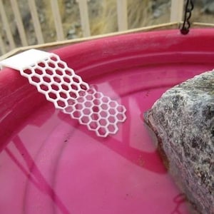 Puede incluir: Una rampa blanca con patrón de panal se extiende hacia una cuenca de agua rosa, junto a una gran roca texturizada. La rampa parece ser un artículo impreso en 3D. El agua es de un color rosa vibrante.