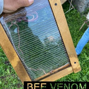 May include: A wooden frame with a clear plastic panel and a wire mesh inside. The frame is brown and has a sign that says "BEE VENOM MACHINE".