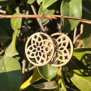 Puede incluir: Un par de pendientes de madera con un diseño geométrico. Los pendientes son de color marrón claro y tienen un aro plateado.