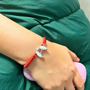 May include: A red braided cord bracelet featuring a central white crescent moon charm. The charm is decorated with a small red square at the center and delicate red dot patterns radiating outward, secured by small red beads on the cord.