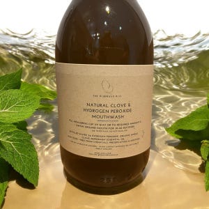 Op de afbeelding: Een bruine glazen fles met een bruin papieren etiket. Het etiket luidt "The Warmann Way Natural Clove & Hydrogen Peroxide Mouthwash Peppermint Flavor". De fles staat op een oppervlak met water en groene bladeren.
