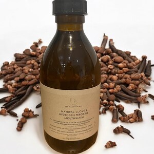 K&ouml;nnte beinhalten: Eine braune Glasflasche mit schwarzem Verschluss, beschriftet mit "The Warmann Way Natural Clove & Hydrogen Peroxide Mouthwash". Die Flasche steht auf einer wei&szlig;en Oberfl&auml;che, im Hintergrund ein Haufen getrockneter Nelken.