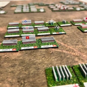 Op de afbeelding: Een close-up van een tabletop-oorlogsspel met miniatuursoldaten en vlaggen. De soldaten zijn in rijen op groene vierkanten gerangschikt met de tekst "Bath's" erop gedrukt. De vlaggen zijn rood, wit en blauw.