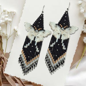 May include: A pair of beaded earrings in a butterfly design. The earrings are black, white, and gold. They are displayed on a white background, with flowers and ribbon.