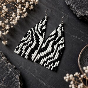May include: A pair of long, triangular dangle earrings crafted from small black and white seed beads arranged in a wavy, abstract pattern. The earrings feature silver-toned metal hooks at the top for attachment, creating a bold, high-contrast design.