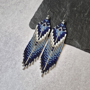 May include: A pair of beaded earrings with a long fringe design. The earrings feature a gradient of blue, white, and gray beads, with silver-colored metal accents. The earrings are displayed on a dark gray surface.