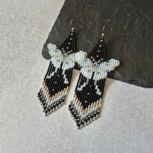 Könnte beinhalten: Ein Paar lange, dreieckige Ohrhänger aus kleinen Glasperlen in einem geometrischen Muster. Das Design besticht durch eine Farbpalette aus Schwarz, Weiß und Metallic-Gold, mit einer spitzen Oberseite und einem gefransten unteren Rand aus Silberperlen.
