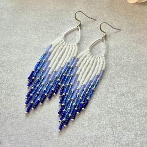 May include: A pair of long, beaded dangle earrings with a gradient of blue and white beads. The earrings are made with small seed beads and have a teardrop-shaped silver hook.