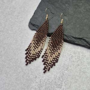 May include: A pair of beaded earrings with a triangular fringe design. The earrings feature a gradient of brown and gold beads, with gold-coloured ear wires. The earrings are displayed on a dark grey surface.