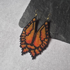 May include: A pair of beaded butterfly wing earrings. The earrings feature a gradient of orange and yellow beads with black bead outlines. Gold-colored hooks are attached to the top of each earring.