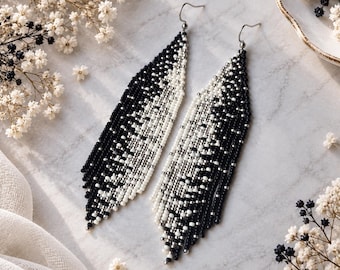 May include: A pair of long, dangling earrings crafted from small, seed-like beads in a gradient pattern of black, white, and silver. The earrings feature a tapered, leaf-like shape with silver-toned metal hooks for attachment to the ear.