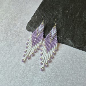 Könnte beinhalten: Ein Paar lange, rautenförmige Ohrhänger aus kleinen Saatperlen in Lavendel, Weiß und Silber. Die Ohrringe zeigen ein geometrisches Muster mit einer Franse aus weißen und lavendelfarbenen Perlen, die an den unteren Kanten herabhängen.