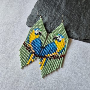 Può includere: Un paio di orecchini pendenti realizzati con piccole perline di forma triangolare. Il design raffigura un pappagallo blu e giallo appollaiato su un ramo marrone su sfondo verde chiaro, con ganci in metallo color argento.