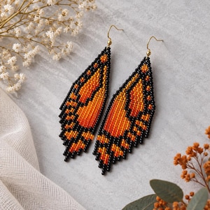 May include: A pair of long, dangling earrings crafted from small glass beads in a vibrant orange and black pattern that mimics the appearance of butterfly wings. The earrings feature gold-toned metal hooks for attachment to the earlobes.