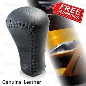 May include: A black leather gear shift knob with visible stitching and the numbers 1, 2, 3, and 4 embossed on the top. The words "Genuine Leather" are printed at the bottom of the image. The knob is designed for a vehicle.