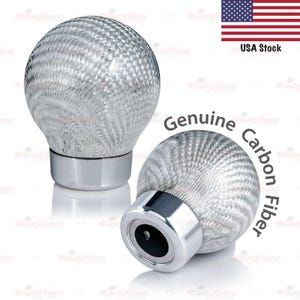 May include: Two silver-colored, spherical car shift knobs with a woven carbon fiber pattern. Each knob has a shiny, chrome-colored base. The text "Genuine Carbon Fiber" is printed on the image.