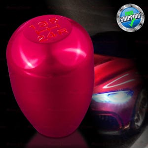 May include: A shiny, magenta-colored car shift knob. The top of the knob has the gear shift pattern in red. The background features a blurred image of a red sports car.