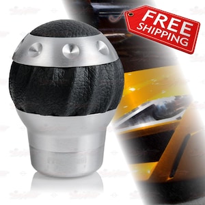 May include: A black leather and silver gear shift knob. The top portion is black leather with a silver band and silver accents. The bottom portion is silver. The gear shift knob is designed for a vehicle.