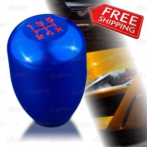 May include: A glossy blue shift knob with red gear markings. The shift pattern is 1, 3, 5, R, 2, and 4. The knob is oval-shaped and appears to be made of metal. The background includes a portion of a yellow car.
