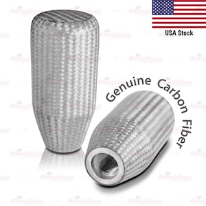 May include: Two silver-colored, carbon fiber gear shift knobs. The knobs have a woven texture and a cylindrical shape. One knob is upright, and the other is lying on its side. The text "Genuine Carbon Fiber" is visible.