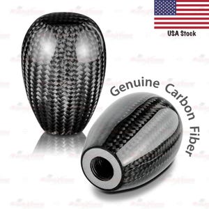 May include: Two black carbon fiber shift knobs. The knobs have a glossy finish and a woven pattern. The text "Genuine Carbon Fiber" is printed on the image. The American flag and the text "USA Stock" are also visible.