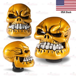 May include: Three gold-colored skull-shaped gear shift knobs with white teeth. The knobs have a metallic finish and are designed with detailed facial features. The image includes a small American flag and the text "USA Stock".