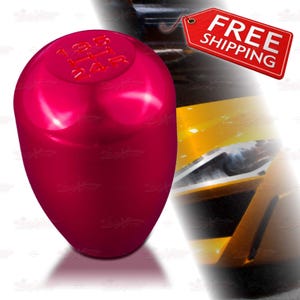May include: A shiny, magenta-colored shift knob with gear markings. The top of the knob has the numbers 1, 2, 3, 4, R. The background includes a partial view of a yellow car.