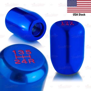May include: Three blue car shift knobs. One is angled, showing the threaded interior. Another shows the top with red gear shift numbers. The third is a side view with red gear shift numbers. The USA flag is in the background.