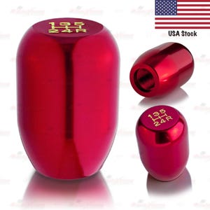 May include: A red, metallic shift knob with gold lettering indicating gear positions. The knob has a smooth, rounded shape and is designed for a vehicle's gear shift lever. The image shows the knob from multiple angles, highlighting its glossy finish.