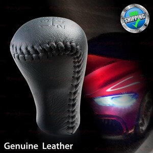 May include: A black leather gear shift knob with white stitching. The numbers 1, 2, 3, 4, and R are visible on the top. The text "Genuine Leather" is at the bottom of the image.