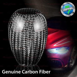 May include: A carbon fiber shift knob with a woven pattern. The knob is oval-shaped and has a glossy finish. The text "Genuine Carbon Fiber" is at the bottom of the image. A red car is in the background.