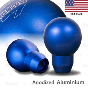 May include: Three blue anodized aluminum shift knobs. The knobs are spherical with a threaded base. The word "MOMO" is visible in white on the top left. The text "Anodized Aluminium" is at the bottom.