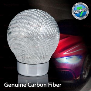 May include: A silver and gray carbon fiber shift knob with a chrome base. The shift knob has a woven texture and is set against a blurred background with the text "Genuine Carbon Fiber".