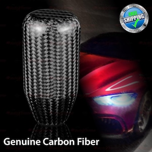 May include: A black carbon fiber shift knob with a woven pattern. The text "Genuine Carbon Fiber" is displayed at the bottom of the image. The background features a red car.