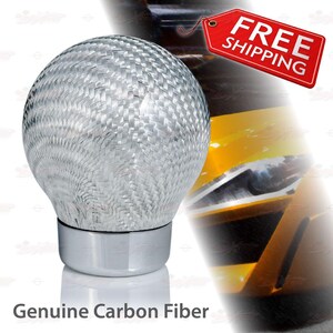 May include: A silver and gray carbon fiber shift knob with a woven texture. The knob has a rounded top and a chrome base. The text "Genuine Carbon Fiber" is displayed below the knob.
